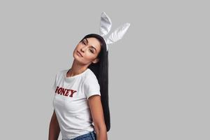 Beautiful bunny. Attractive young woman in bunny ears looking at camera and smiling while standing against grey background photo