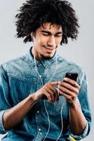 Creating new playlist. Cheerful young African man wearing headphones and using smartphone with smile while sitting against grey background photo