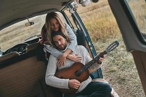 tocando una canción romántica. un joven apuesto tocando la guitarra para su bella novia mientras se sienta en una mini furgoneta de estilo retro foto