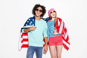despreocupado y enamorado. funky pareja joven que cubre con la bandera americana y sonriendo mientras está de pie contra el fondo blanco foto