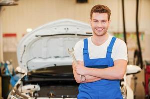 He is ready to work. Confident young man in uniform holding a wrench an smiling while standing in workshop with car in the background photo