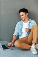 Time to relax. Smiling young man working on laptop and holding cup of coffee while sitting against grey background photo