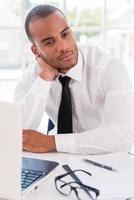 Lost in business thoughts. Thoughtful young African man in shirt and tie leaning his head on hand and looking away while sitting at his working place photo