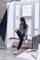 Writing down ideas. Thoughtful young woman writing something in her notebook while sitting on the window sill photo