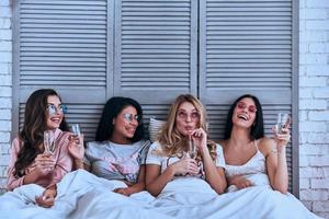 divirtiéndose. cuatro atractivas mujeres jóvenes en pijama bebiendo cócteles y sonriendo mientras están en la cama foto