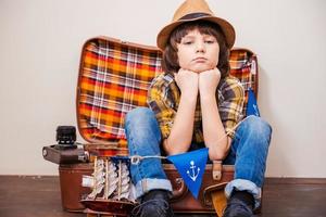 Tired from adventures. Little boy in headwear holding hands on chin and looking at camera while sitting on suitcase against brown background photo