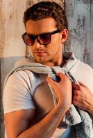 Making his style. Handsome young man in sunglasses adjusting his shirt while standing against the wooden wall photo