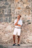 Dreaming about something good. Full length of cheerful young African man in smart casual clothes holding hand on chin and looking thoughtful while standing against the stoned wall outdoors photo