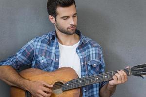 Music is my life. Handsome young man playing the guitar while standing against grey background photo