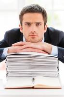 Feeling exhausted. Depressed young man in formalwear looking at camera and leaning his face at the stack of documents laying on the table photo