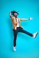 Full length of playful young African man wearing virtual reality glasses while hovering against blue background photo