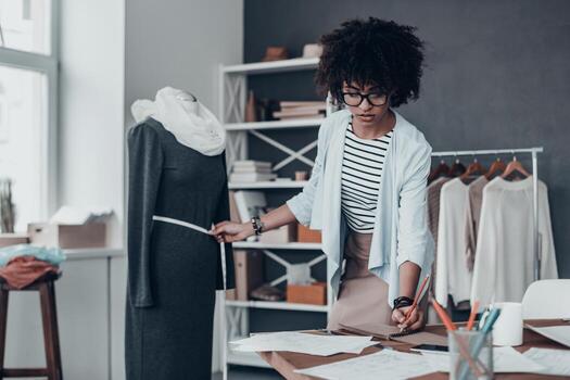 trabajando en nueva colección. joven africana concentrada midiendo la cintura con cinta adhesiva y anotando los resultados mientras está de pie en su taller con ropa colgada en el fondo foto
