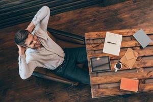 Satisfied with his work. Top view of handsome young man keeping hands behind head and smiling while sitting at his working place photo