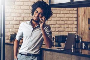 We are open today Cheerful young African man talking on mobile phone with smile and looking away while standing at bar counter photo