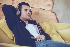 Great song Young man in headphones keeping eyes closed while sitting on couch at home photo