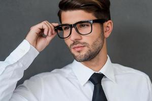 Confident and handsome. Confident young businessman adjusting his eyeglasses and looking away while standing against grey background photo