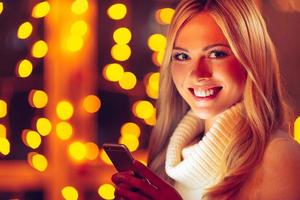 Always in touch with her nearest. Beautiful young smiling woman in white sweater holding smart phone and looking at camera with defocused Christmas lights in the background photo