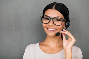 Service with happy smile. Cheerful young female customer service looking at camera and smiling while standing against grey background photo