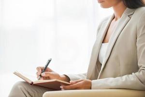 Making business notes. Side view of young businesswoman in suit writing something in note pad while sitting at the chair photo