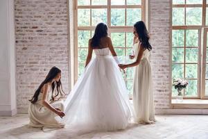 comienzo del día más feliz. Longitud total de damas de honor ayudando a la novia con su vestido de novia mientras están juntas cerca de la ventana foto