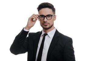 Feeling confident. Handsome young man in full suit adjusting his eyeglasses and looking at camera while standing against white background photo