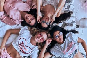 Hurry to be happy. Top view of four playful young women in pajamas and eyewear smiling while lying on the bed at home photo