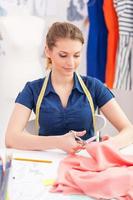 Confident tailor at work. Beautiful young woman with measuring tape on shoulders cutting the cloth with scissors while sitting at her working place photo