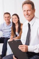 Confident financial expert. Confident mature man in shirt and tie holding clipboard and looking at camera while couple sitting in the background and smiling photo