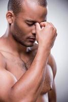 estresado y cansado. retrato de un hombre africano sin camisa tocándose la nariz y manteniendo los ojos cerrados mientras se enfrenta a un fondo gris foto