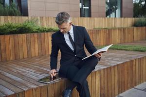 hombre de negocios maduro confiado que usa una tableta digital y toma notas mientras se sienta al aire libre cerca del edificio de oficinas foto