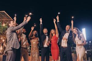 Group of beautiful people in formalwear holding sparklers and smiling while spending time on outdoor party photo