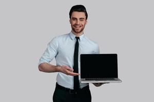 Copy space on his laptop.  Good looking young man in white shirt and tie pointing copy space on his laptop and smiling while standing against grey background photo