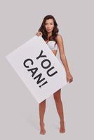 Everyone needs a bit of encouragement.  Full length of attractive young woman holding a poster and looking at camera while standing against grey background photo