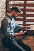 Coffee break. Side view of handsome thoughtful young man holding smart phone and looking at it while sitting near window in loft interior with coffee cup in his hand photo