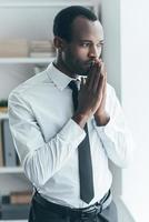 Thinking about new project. Handsome young African man holding hand clasped near face and looking away while standing in creative office photo