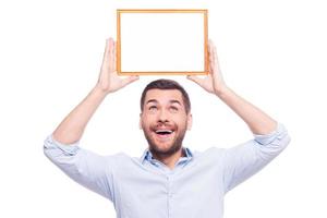 Happy about that... Handsome young man in shirt holding a picture frame upon his head and looking up on it while standing against white background photo