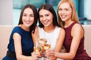 disfrutando de un gran tiempo juntos. tres hermosas mujeres jóvenes en traje de noche sentadas en el sofá y sosteniendo copas con vino foto