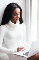 Surfing the net at home. Attractive young African woman in warm sweater working on laptop while sitting on the window sill photo