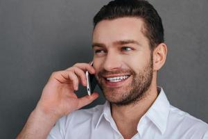 Good talk. Handsome young man talking on the mobile phone and smiling while standing against grey background photo