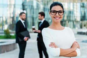 Confident businesswoman. Smiling young businesswoman keeping arms crossed and looking at camera while two her male colleagues talking to each other in the background photo