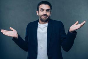 Who knows Confused young man stretching out his arms and grimacing while standing against grey background photo