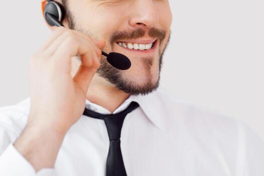 ¿Cómo puedo ayudarte? imagen recortada de un joven apuesto con ropa formal ajustando sus auriculares y sonriendo mientras está de pie contra un fondo gris foto
