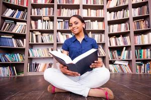 le encanta leer. hermosa estudiante africana sosteniendo un libro y sonriendo a la cámara mientras se sienta en el suelo en la biblioteca foto