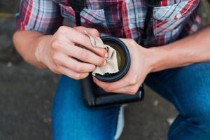 lente de limpieza del fotógrafo. primer plano del hombre limpiando la lente de su cámara digital mientras está de pie al aire libre foto