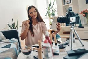 mujer joven segura de sí misma que aplica un pincel de maquillaje y sonríe mientras hace un video en las redes sociales foto