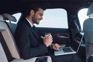 Driven by success. Handsome young man in full suit working using laptop while sitting in the car photo
