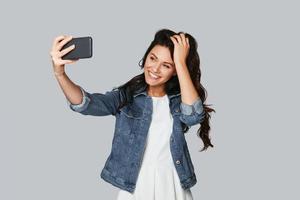 Sharing nice photo. Attractive young woman smiling and taking selfie while standing against grey background photo