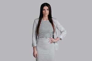 Confident in her style.  Beautiful young woman in smart casual wear keeping hand on hip and looking at camera while standing against grey background photo