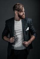 Confident handsome. Young hipster looking away and adjusting his jacket while standing against grey background photo