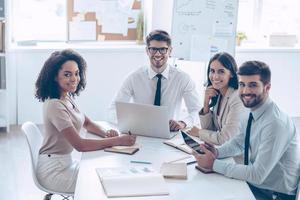 Best business team ever Group of cheerful coworkers looking at camera with smile while sitting at the office table photo